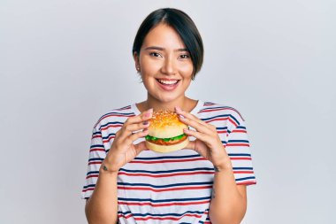 Kısa saçlı, hamburger yiyen, yüzünde mutlu ve havalı bir gülümsemeyle gülümseyen güzel bir genç kadın. diş gösterme. 