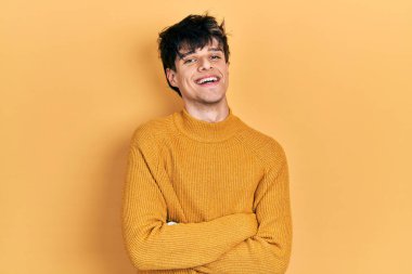 Yakışıklı hippi genç adam gündelik sarı süveter giyip kollarını kavuşturup kameraya bakarken gülümsüyor. pozitif kişi. 