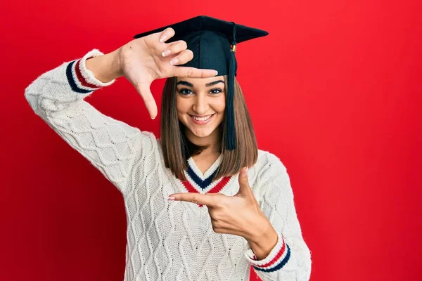 卒業キャップを身に着けている若いブルネットの女の子は幸せな顔で手と指でフレームを作り笑顔 創造性と写真の概念 — ストック写真