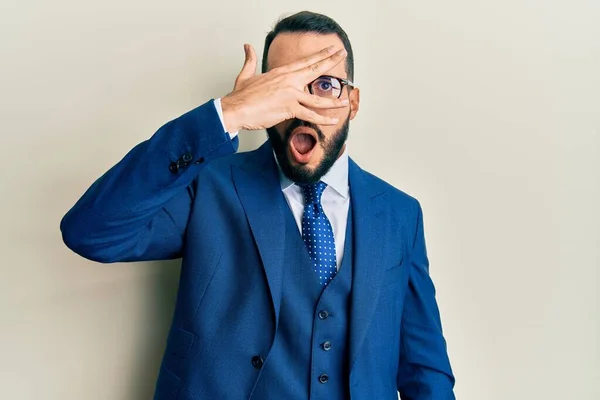 Young man with beard wearing business suit and tie peeking in shock covering face and eyes with hand, looking through fingers with embarrassed expression.