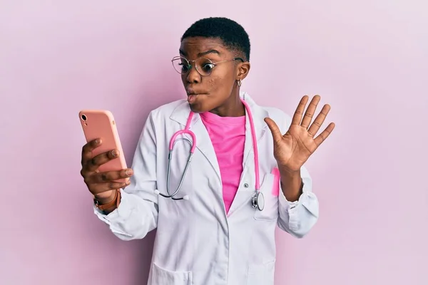 Young african american doctor woman doing video call using smartphone making fish face with mouth and squinting eyes, crazy and comical.