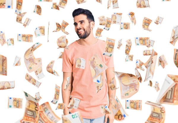 Молодой красивый мужчина с бородой в повседневной футболке смотрит в сторону с улыбкой на лице, естественным выражением лица. смеясь уверенно.