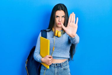 Genç esmer kız öğrenci çantası ve kulaklık takıyor. El açık, dur işareti yapıyor. Ciddi ve kendinden emin bir ifadeyle, savunma jesti. 