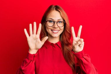 Genç, güzel, kızıl saçlı bir kadın kırmızı arka planda günlük kıyafetler ve gözlük takıyor ve yedinci parmaklarıyla işaret ederken kendinden emin ve mutlu bir şekilde gülümsüyor.. 