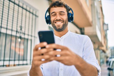 Güneşli bir günde sakallı yakışıklı bir adam açık havada mutlu bir şekilde gülümsüyor kulaklık takarak müzik dinliyor.