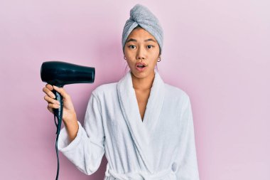 Duş havlusu şapkası ve bornoz giyen genç Çinli kadın kurutucuyu tutarken korkmuş ve ağzı açık kalmış şaşkınlık içinde, inançsız bir yüz. 