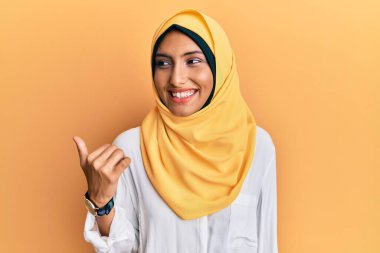 Genç esmer Arap kadın geleneksel İslami başörtüsü takıyor mutlu yüzle gülümsüyor ve başparmağıyla yana bakıyor.. 