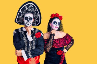 Young couple wearing mexican day of the dead costume over background looking confident at the camera smiling with crossed arms and hand raised on chin. thinking positive. 
