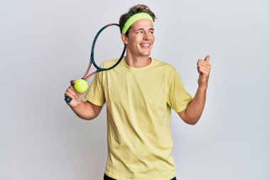 Tenis oynayan yakışıklı beyaz adam raket ve top tutuyor başparmağıyla yan tarafa bakıyor ve ağzı açık gülümsüyor. 