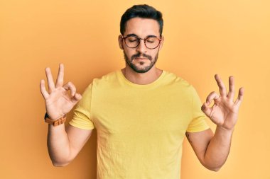 Gündelik kıyafetler giyen, gözlüklü, gülümseyen, gözleri kapalı meditasyon yapan genç İspanyol adam parmaklarıyla meditasyon yapıyor. yoga konsepti. 