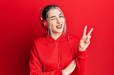 Spor kıyafetleri giyen, kulaklık takan, kameraya gülümseyen ve zafer işareti yapan genç modern bir kız. İki numara.. 