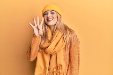 Genç beyaz kadın yün kazak ve kep giyip dört numaralı parmaklarıyla işaret ederken kendinden emin ve mutlu bir şekilde gülümsüyor.. 