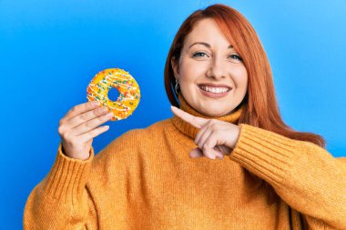 Güzel kızıl saçlı kadın elinde lezzetli renkli donut tutarken gülümsüyor mutlu bir şekilde eli ve parmağı ile işaret ediyor. 