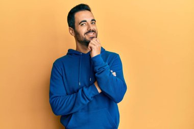 Gündelik kıyafetler giyen genç İspanyol adam kameraya bakıyor kollarını kavuşturup gülümseyerek ve çenesini kaldırarak. Olumlu düşünmek. 