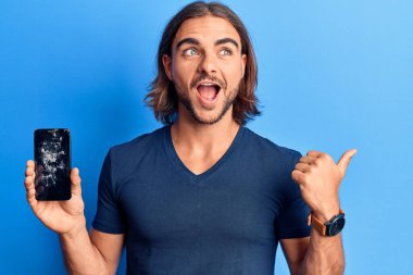 Kırık akıllı telefon tutan genç yakışıklı adam kırık ekranı gösteriyor başparmağıyla yan tarafa bakıyor mutlu bir şekilde gülümsüyor. 