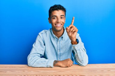 Genç, yakışıklı, Afro-Amerikalı, gündelik giysiler giymiş masada oturmuş başarılı bir fikirle işaret ediyor. Heyecanlı ve mutlu. Bir numara.. 