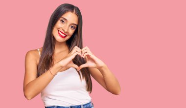 Gündelik giysiler giyen genç İspanyol kadın. Sevgiyle gülümsüyor. Kalp sembolü ve şekli elleriyle gösteriyor. romantik kavram. 