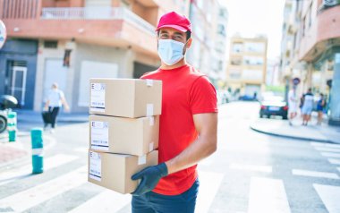 Üniforma giyen ve Coronavirus koruması takan genç bir teslimatçı. Sağlık maskesi. Kasaba sokaklarında karton paketler tutuyor..