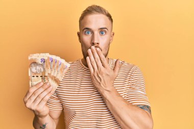 Güney Afrika 'da elinde 20 rand banknot tutan genç beyaz bir adam ağzını eliyle kapatıyor, şok geçiriyor ve hata yapmaktan korkuyor. şaşırmış ifade 