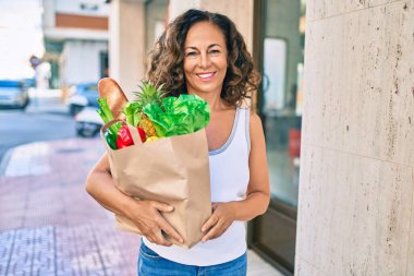 Orta yaşlı İspanyol kadın, elinde alışveriş torbalarıyla şehirde mutlu bir şekilde gülümsüyor..