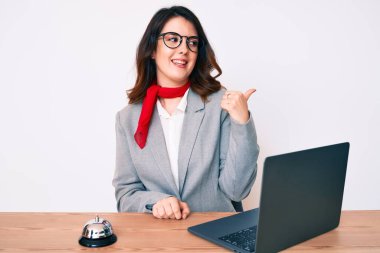 Genç güzel esmer kadın otel resepsiyonunda çalışırken dizüstü bilgisayarla gülümsüyor, gülümsüyor ve başparmağıyla yan tarafı işaret ediyor.. 