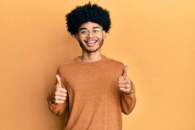 Afro saçlı, kışlık süveter giyen, elinde olumlu jest yapan, gülümseyen ve mutlu genç Afro-Amerikalı bir adam. Neşeli ifade ve kazanan jest. 