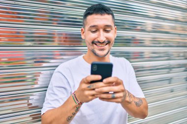 Duvara dayanmış akıllı telefon kullanan, mutlu gülümseyen genç İspanyol adam.