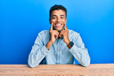 Genç, yakışıklı, Afro-Amerikalı, gündelik giysiler içinde masada oturmuş ağzı açık gülümsüyor, parmakları işaret ediyor ve zorla neşeli bir gülümseme sergiliyor. 