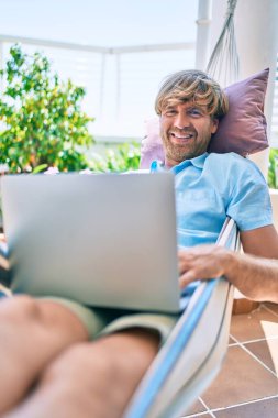 Evinin terasındaki orta yaşlı yakışıklı adam hamakta uzanıp dizüstü bilgisayarla çalışıyor.