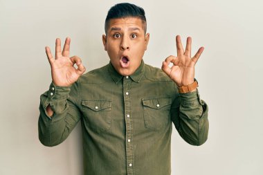 Gündelik kıyafetler giyen genç latin adam şaşırmış ve şok olmuş görünüyor tamam onay sembolünü parmaklarıyla yapıyor. çılgın ifade 