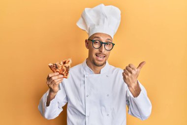 Kel, sakallı, profesyonel aşçı önlüğü giymiş, elinde İtalyan pizzası tutan, baş parmağını yana doğru gösteren, ağzı açık, mutlu bir şekilde gülümseyen bir adam. 