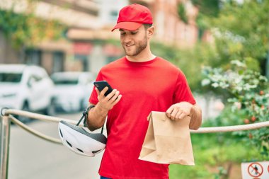 Beyaz atlı genç teslimatçı akıllı telefon kullanıyor ve şehirde kağıt torba taşıyor..