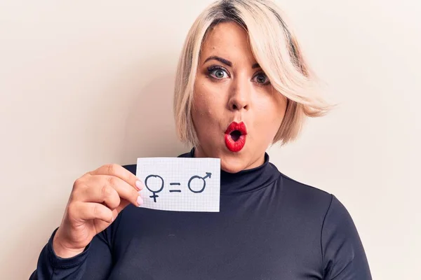 Blonde plus size woman asking for sex equality holding paper with gender equal message scared and amazed with open mouth for surprise, disbelief face