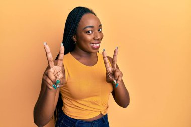 Gündelik giysiler giymiş genç Afro-Amerikan kadın kameraya bakıp zafer işareti yapıyor. İki numara.. 