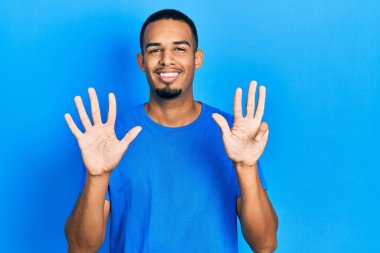 Genç Afro-Amerikan adam günlük mavi tişört giyer ve dokuz numaralı parmaklarıyla işaret ederken kendinden emin ve mutlu gülümser.. 