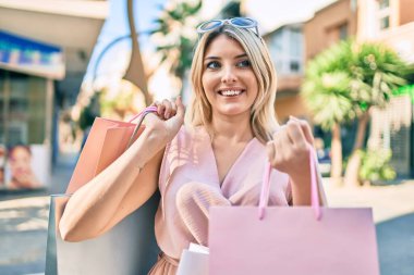 Genç sarışın kadın gülümsüyor. Alışveriş torbalarını şehirde tutarken mutlu..