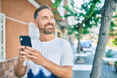 Orta yaşlı yakışıklı adam şehirde akıllı telefon kullanarak mutlu gülümsüyor..