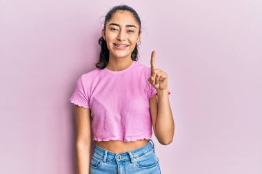 Diş teli takan, sıradan kıyafetler giyen, bir fikir veya soruyla gülümseyen, mutlu bir yüzle işaret eden İspanyol bir genç kız. 