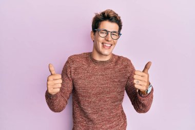 Yakışıklı beyaz adam, günlük süveter ve gözlük takıyor, başarı işareti yapıyor, el hareketi yapıyor, gülümsüyor ve mutlu. Neşeli ifade ve kazanan jest. 