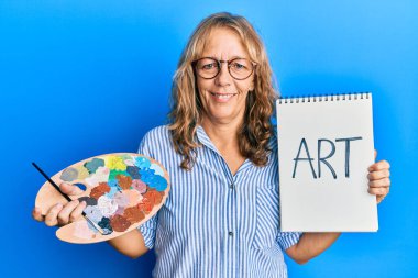 Orta yaşlı sarışın kadın elinde ressam paleti ve defteriyle yüzünde mutlu ve havalı bir gülümsemeyle gülümsüyor. diş gösterme. 