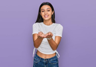 Elleri avuçlarında gülümseyen ya da el hareketi yapan günlük beyaz tişört giyen İspanyol genç kız. Bekleme ve koruma 