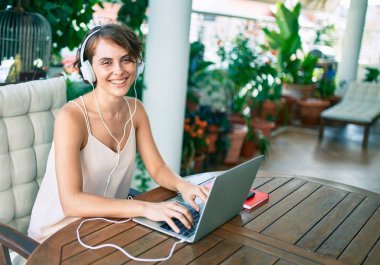 Genç, güzel, beyaz bir kadın mutlu bir şekilde gülümsüyor. Evde çalışıyor ve kulaklık takarak müzik dinliyor.