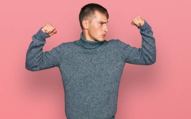 Sıradan kıyafetler giyen genç sarışın adam gururla gülümseyen kol kaslarını gösteriyor. fitness konsepti. 