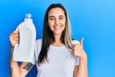 Deterjan şişesi tutan genç İspanyol kadın mutlu ve pozitif gülümsüyor, başparmağıyla mükemmel ve onay işareti yapıyor. 