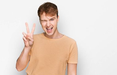 Gündelik kıyafetler giyen, kameraya gülümseyen ve parmaklarıyla zafer işareti yapan beyaz bir genç. İki numara.. 