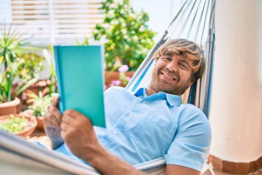 Orta yaşlı yakışıklı bir adam evinin terasında bir hamakta uzanıp kitap okuyor.