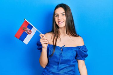 Sırbistanlı bayrak tutan genç esmer kız pozitif ve mutlu görünüyor ve kendine güvenen bir gülümsemeyle dişlerini gösteriyor. 