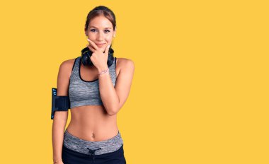 Spor kıyafetleri giyen ve kulaklık takan genç ve güzel bir İspanyol kadın kameraya kendinden emin bir şekilde bakıyor kollarını kavuşturmuş ve çenesini kaldırmış gülümsüyor. Olumlu düşünmek. 