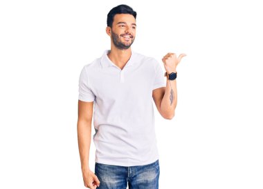 Genç, yakışıklı, günlük kıyafetler giyen mutlu yüzlü gülümseyen ve başparmağıyla yan tarafı gösteren İspanyol bir adam.. 