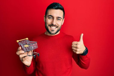 Genç İspanyol adam elinde küçük bir süpermarket arabası tutuyor mutlu ve pozitif gülümsüyor, başparmağıyla mükemmel ve onay işareti yapıyor. 